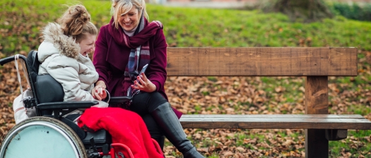 A person with disability sits in a wheelchair, smiling with their support person in a park
